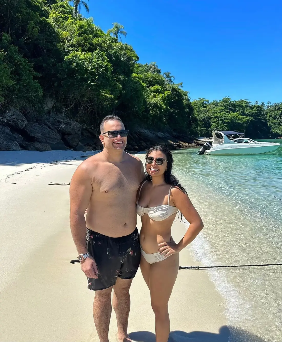 Casal na areia de uma praia com a lancha ancorada ao fundo